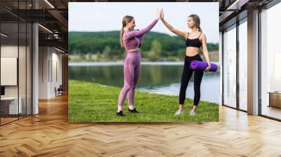 Two girls give each other high five after a good training session in the park near lake. Attractive sporty women smiling outdoor. Wall mural